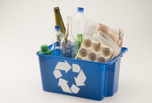 A blue recycling bin filled with various types of waste materials, including several clear and green plastic bottles with screw caps, a cluster of transparent glass bottles with narrow necks, a cardboard egg carton, and crumpled paper. The bottles vary in size and shape, with some standing upright and others leaning against the sides of the bin. The cardboard egg carton appears used, with some eggshell fragments or residue visible inside. The bin's surface displays a white recycling symbol composed of three arrows forming a triangle, indicating its use for recyclable rubbish. The background is plain white, and the lighting is even, highlighting the textures and colors of the materials. This scene exemplifies typical waste collection for rubbish removal services, such as those provided by House Clearance Purfleet, focusing on domestic recyclable waste.