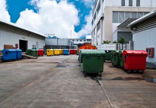 A concrete outdoor yard area between two industrial or commercial buildings, featuring various large wheeled waste disposal bins in multiple colours including green, red, yellow, and blue, arranged along the sides and towards the background. The bins are made of plastic with textured surfaces and fitted with lids, some of which are closed while others are open or leaning. The surrounding buildings have metal cladding and large garage doors, with the left building painted in light grey and the right in a similar tone, with additional external ventilation. The ground surface is paved with concrete slabs, showing minor stains and cracks. In the background, a clear blue sky with some scattered white clouds is visible, indicating daylight. This scene illustrates typical rubbish collection or waste management setup, relevant to local household or commercial clearance services in Purfleet or nearby areas, as managed by House Clearance Purfleet, offering professional rubbish removal solutions.