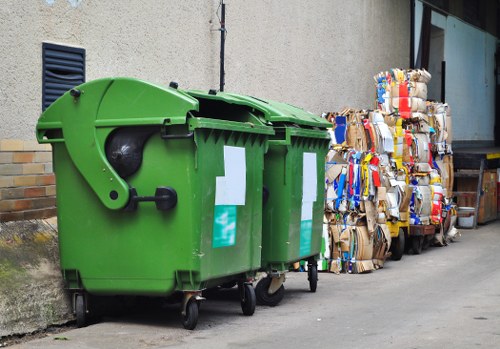 A large green wheeled rubbish bin with a black lid, positioned on a paved driveway against a beige exterior wall of a property. The bin has a matte surface with some white labels attached to its side. To the right of the bin, there are neatly stacked bundles of flattened cardboard boxes and paper, tied together with string, showing a mix of brown, white, and coloured packaging materials. In the background, part of a window with reflective glass and a wooden pallet are visible, indicating a residential or commercial premises. The scene suggests a collection or removal of waste materials near a home or business location, consistent with waste management services provided by House Clearance Purfleet, especially within the Purfleet area.