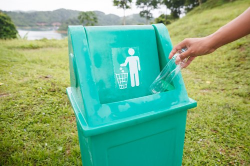A teal-colored outdoor waste bin with a white icon depicting a person discarding rubbish into a bin, situated on a grassy area with a lake and trees in the background. A person's hand is seen disposing of a clear plastic container into the bin, which is positioned on a paved or grassy verge near a natural landscape. The scene suggests a clean, environmentally conscious setting, and the image relates to waste disposal services offered by House Clearance Purfleet, supporting rubbish removal in residential or natural environments within the local area including Purfleet in Essex.