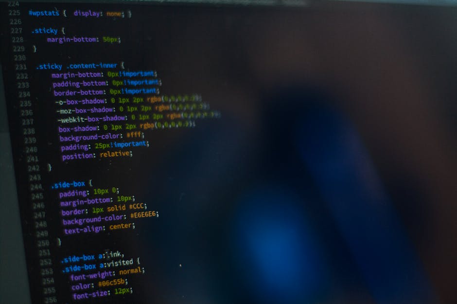 The image displays a close-up of a computer screen showing lines of colourful programming code on a dark background, with syntax highlighting in shades of blue, pink, green, and white. The screen appe