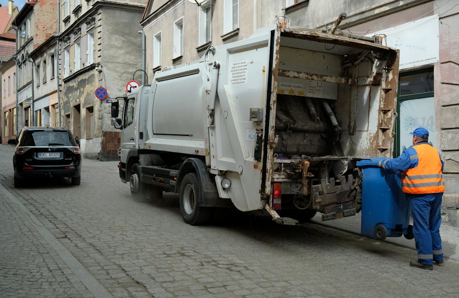 A roadside scene features a municipal waste collection vehicle with a primarily red exterior, positioned on a street beside a curb. The vehicle's large rear hopper is open, revealing mechanical compon