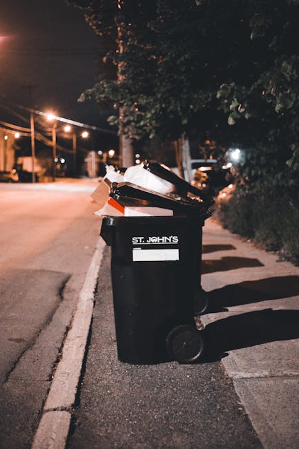 Two large black plastic rubbish bags, appearing crumpled and slightly torn, are placed on the pavement near the curb in an outdoor urban setting. The bags are filled with waste and are positioned clos
