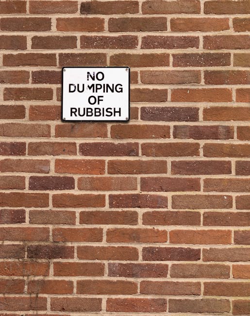 A rectangular black and white metal sign affixed to a red and brown brick wall displaying the words 'NO DUMPING OF RUBBISH.' The bricks are arranged in a horizontal running bond pattern, with some bri